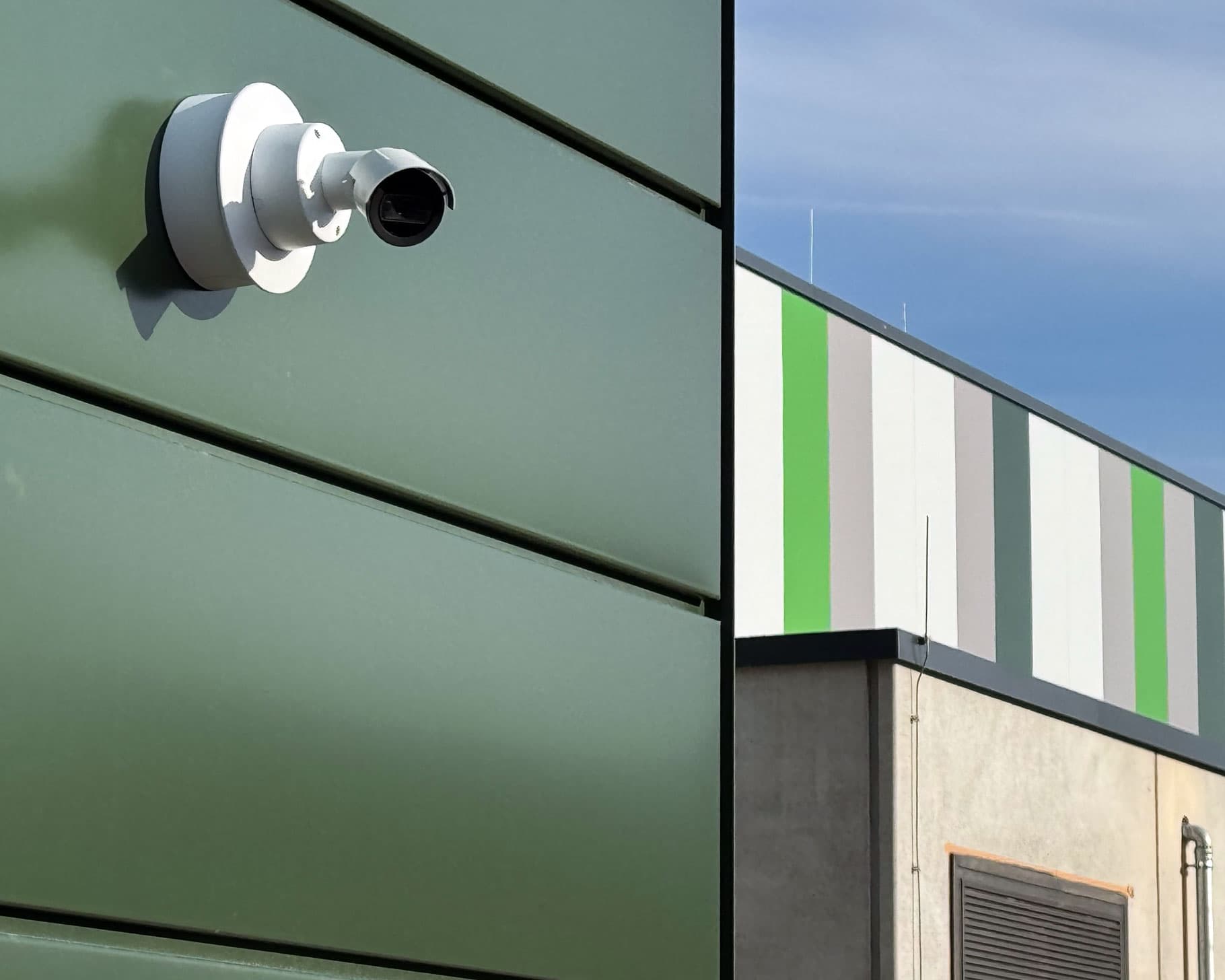 Weiße Bullet-Kamera an einer grünen Metallfassade eines Gewerbegebäudes vor blauem Himmel.